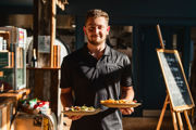 A young man in a black uniform is carrying two plates of food from the kitchen. There is a chalkboard to his left.