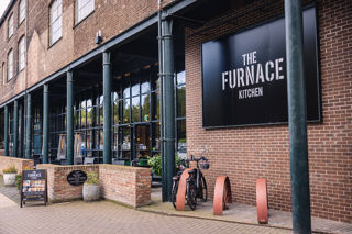 Modern brick wall and glass fronted cafe building with a large black sign with white writing saying The Furnace Kitchen.